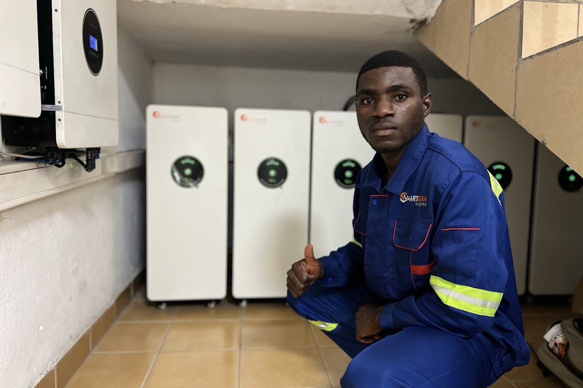 A technician in SmartSana Energies overalls crouching beside a row of battery cabinets and a hybrid inverter
