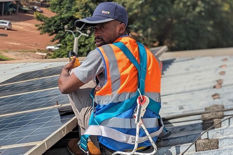 A technician in a safety harness and high-visibility vest checking solar panels on a roof with a clamp meter