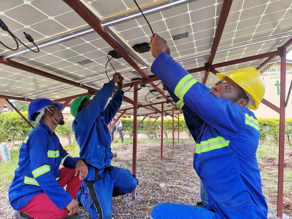 Trainees in hard hats and high-visibility overalls wiring solar modules under a ground-mounted array
