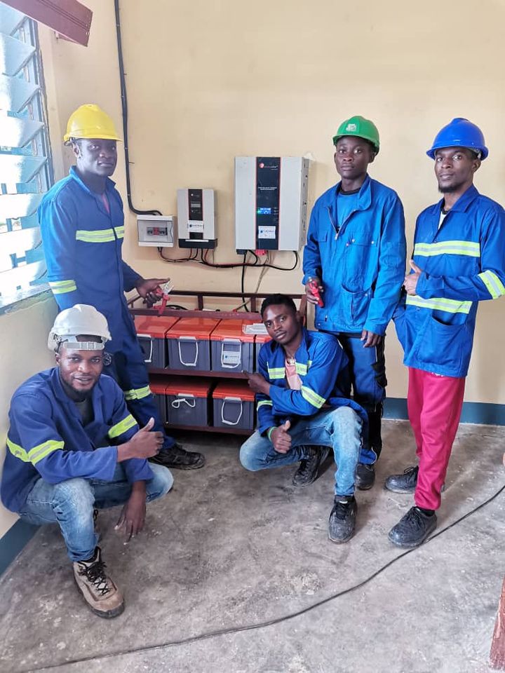 Five trainees beside a finished installation: inverters, a protection box and a battery bank